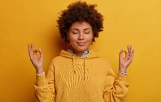 Portrait of ethnic woman stays calm, meditates and practices yoga, keeps hands in zen gesture, closes eyes, relaxes after hard working day, wears yellow sweatshirt, poses indoor, unites with nature