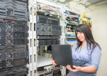The girl programmer stands near the racks with computer equipment. System administrator to configure network protocols. A woman with a laptop works in a server room.