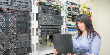 The girl programmer stands near the racks with computer equipment. System administrator to configure network protocols. A woman with a laptop works in a server room.