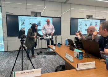 A group of technology professionals participates in a live Tech Field Day X session at HPE’s Houston campus. Two presenters, a woman and a man, stand at the front demonstrating the internal hardware of an HPE ProLiant DL145 Gen11 server. The background displays slides highlighting the server's key features. Around the table, attendees including Keith Townsend, Stephen Foskett, and Guy Currier are engaged—some taking notes, others capturing the demo on their phones. A camera on a tripod records the session. Nameplates and product boxes are visible on the conference table.