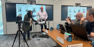 A group of technology professionals participates in a live Tech Field Day X session at HPE’s Houston campus. Two presenters, a woman and a man, stand at the front demonstrating the internal hardware of an HPE ProLiant DL145 Gen11 server. The background displays slides highlighting the server's key features. Around the table, attendees including Keith Townsend, Stephen Foskett, and Guy Currier are engaged—some taking notes, others capturing the demo on their phones. A camera on a tripod records the session. Nameplates and product boxes are visible on the conference table.