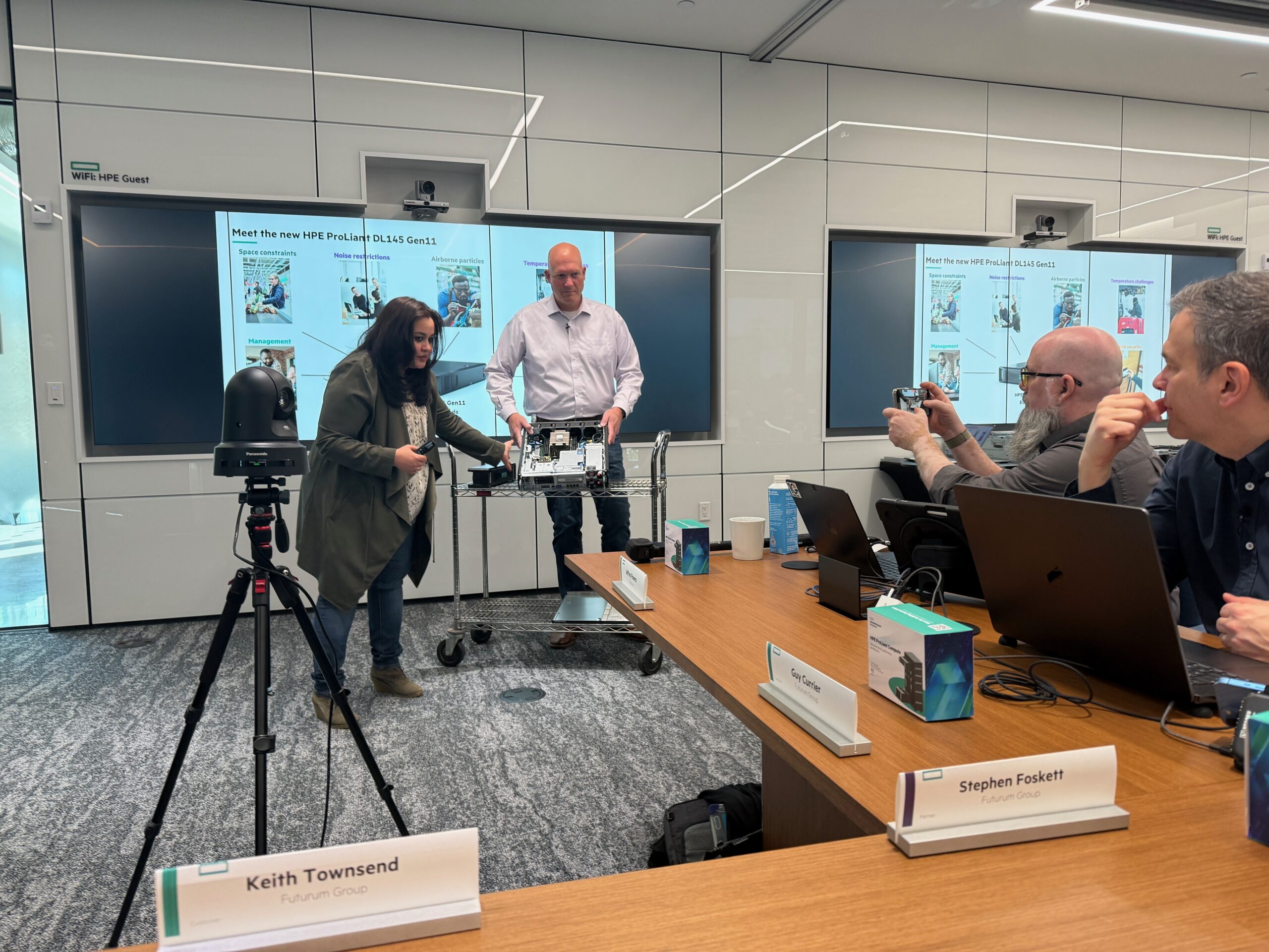 A group of technology professionals participates in a live Tech Field Day X session at HPE’s Houston campus. Two presenters, a woman and a man, stand at the front demonstrating the internal hardware of an HPE ProLiant DL145 Gen11 server. The background displays slides highlighting the server's key features. Around the table, attendees including Keith Townsend, Stephen Foskett, and Guy Currier are engaged—some taking notes, others capturing the demo on their phones. A camera on a tripod records the session. Nameplates and product boxes are visible on the conference table.